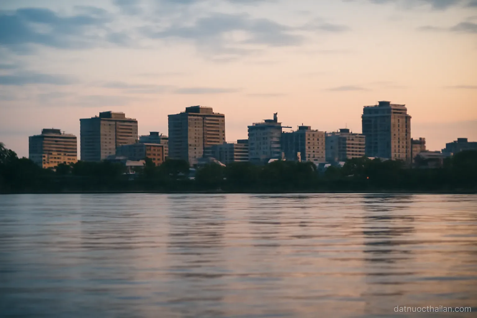 A city skyline of mid-rise buildings seen across a wide river at dusk, lights not yet on